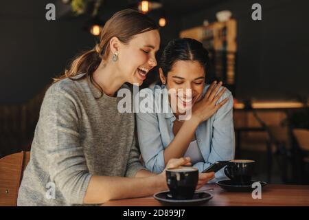Frau, die ihrem Freund lustige Posts auf ihrem Handy zeigt und lächelt. Zwei Freundinnen sitzen im Coffeeshop mit einem Handy und lächeln. Stockfoto