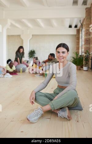 Porträt einer jungen gesunden Frau, die beim Sitzen die Kamera anlächelt Auf dem Boden mit anderen Personen im Hintergrund Tanzstudio Stockfoto