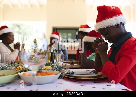 Multigeneration Familie trägt santa Hüte beten, bevor sie gemeinsam Mittagessen, während auf dem Esstisch im Wohnzimmer zu Hause sitzen. weihnachten fes Stockfoto