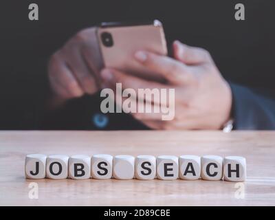 Closeup Holzblock schriftliche Jobs Suche auf dem Tisch gegen bussinessman mit Smartphone, um neue Jobs und Anwendung zu finden. Beschäftigungskonzept. Stockfoto