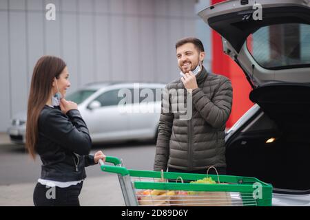 Glücklicher Mann und seine Frau ziehen Schutzmaske Outddors nach dem Einkaufen in Lebensmitteln oder Supermarkt während Quarantäne. Trolley voll mit frischen Lebensmitteln Stockfoto