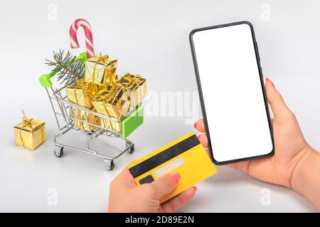 Frau benutzt Handy und Kreditkarte Mockup, um Weihnachtseinkäufe zu machen. Hintergrund der kleinen Supermarkt Warenkorb mit Weihnachtsgeschenken gefüllt. Online-Cris Stockfoto