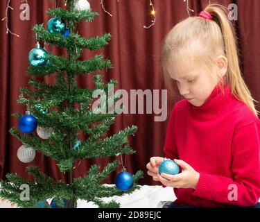 Ein niedliches kaukasisches fröhliches Mädchen in einem roten Pullover hält eine blaue Kugel in den Händen, um an einem dekorierten künstlichen Weihnachtsbaum zu hängen, schmückt das Kind t Stockfoto