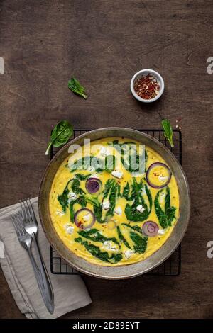 Hausgemachte, leckere Omelette oder Frittata mit Spinat, Feta-Käse, rote Zwiebeln in einer Eisenpfanne auf einem rustikalen Tisch. Draufsicht Stockfoto