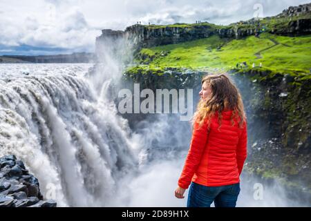 Junge Frau zurück Blick auf Dettifoss Wasserfall auf Felsen Wasser Fließender Nebel versprüht bewölkten Tag in Island mit oranger Jacke Und Jeans Stockfoto