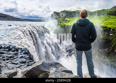 Junger Mann zurück Blick auf Dettifoss Wasserfall auf Felsen Wasser Fließender Nebel versprüht bewölkten Tag in Island mit schwarzem Mantel Stockfoto