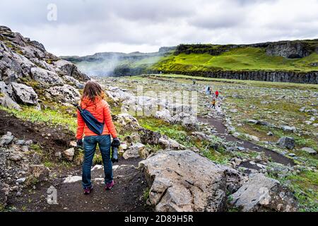 Junge Frau wieder stehend Blick auf Dettifoss Wasserfall auf Felsen Wassernebel versprüht bewölkten Tag in Island mit oranger Jacke Und Jeans Stockfoto