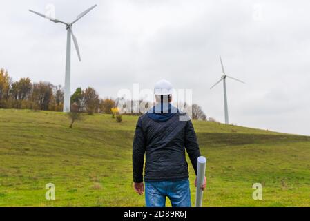 Rückansicht eines männlichen Ingenieurs trägt weißen Helm, der hält Ein Plan am Standort der Windturbine Herbst wolkig Himmel grün Wiese Stockfoto