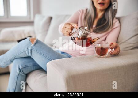 Frau mittleren Alters sitzt auf der Couch und gießt Tee in die Tasse Stockfoto