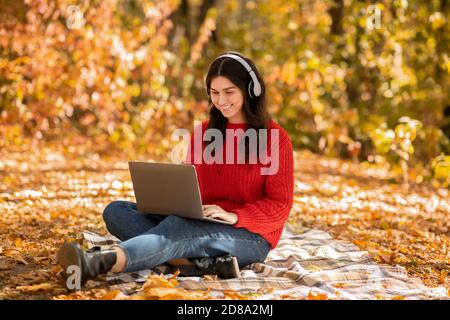 Porträt einer Studentin in voller Länge mit Laptop und Kopfhörer Teilnahme an Online-Unterricht im Herbst Park Stockfoto