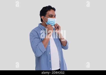 Indischer Mann setzt auf medizinische Maske isoliert. Mann in blauem Hemd zeigt, wie man auf medizinische Maske setzen. Gesundheit, Virus, Medizinisches Konzept Stockfoto