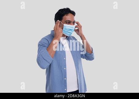 Indischer Mann setzt auf medizinische Maske isoliert. Mann in blauem Hemd zeigt, wie man auf medizinische Maske setzen. Gesundheit, Virus, Medizinisches Konzept Stockfoto