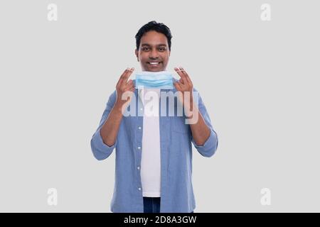 Indischer Mann setzt auf medizinische Maske isoliert. Mann in blauem Hemd zeigt, wie man auf medizinische Maske setzen. Gesundheit, Virus, Medizinisches Konzept Stockfoto