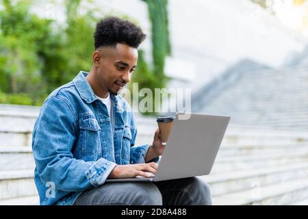 Porträt des jungen afrikanischen Mann in Jeans Jacke arbeiten auf Laptop im Freien, Papier Kaffee Tasse o Stein Sitz in seiner Nähe Stockfoto