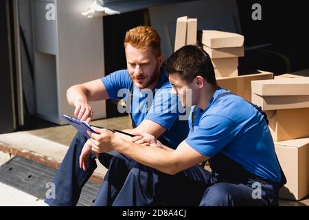 Movers in Overalls Blick auf Zwischenablage in der Nähe von Kartons in Lager Stockfoto