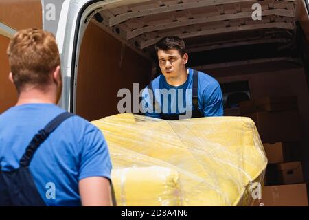 Selektiver Fokus des Laders, der die Couch in Stretch-Wrap trägt Mit Mitarbeiter in der Nähe von LKW mit offenen Türen im Freien Stockfoto