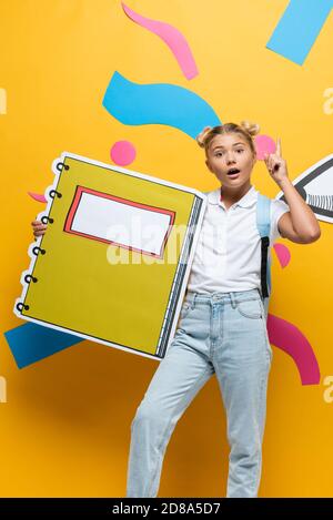 Stauned Schülerin mit Notebook-Maquette zeigt Idee Geste auf gelb Hintergrund mit bunten Elementen und Papier geschnitten Bleistift Stockfoto