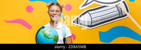 Horizontales Bild von Schulkind mit Globus Blick auf Kamera in der Nähe Papierhandwerk auf gelbem Hintergrund Stockfoto