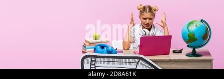 Horizontale Ernte von aggressiven Mädchen in Kopfhörer sitzen in der Nähe von Laptop, Bücher und Papierkunst isoliert auf rosa Stockfoto