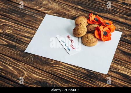 Karte mit anzas Tagesschriftzug bei Kunstblumen, Keksen und leerem Papier auf Holzfläche Stockfoto