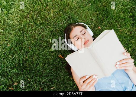 Draufsicht auf attraktive Frau in schnurlosen Kopfhörern liegend Gras und Buch lesen Stockfoto
