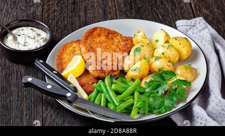 Deutsches Schweinsschnitzel mit jungen Kartoffeln, grüne Bohnen mit Besteck auf einem Teller mit Zitronenkeilen serviert, und Mayonnaise auf Basis Sauce auf einem dunklen Holz b Stockfoto