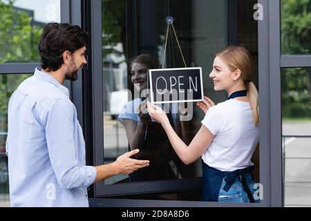 Der Kunde zeigt mit der Hand in der Nähe der lächelnden Kellnerin und hält das Schild mit Öffnen Sie die Beschriftung an der Tür des Cafés Stockfoto