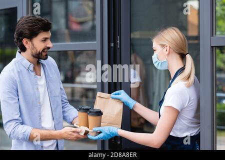 Seitenansicht des Mannes, der das Paket und die Pappbecher von empfängt Kellnerin in Latexhandschuhen und medizinischer Maske in der Nähe des Cafés Stockfoto