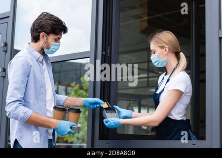 Seitenansicht des Mannes, der für die Bestellung mit Kreditkarte bezahlt Und Verkäuferin mit Zahlungsterminal in der Nähe von Café Stockfoto