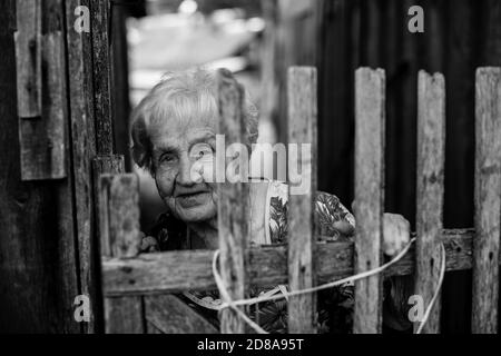 Eine ältere Frau schaut hinter einem Zaun im Dorf hinaus. Schwarzweiß-Fotografie. Stockfoto