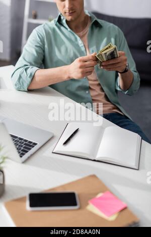 Beschnittene Ansicht des Mannes, der Bargeld in der Nähe von digitalen Geräten und Notebooks auf dem Tisch zu Hause hält, verdienen Online-Konzept Stockfoto