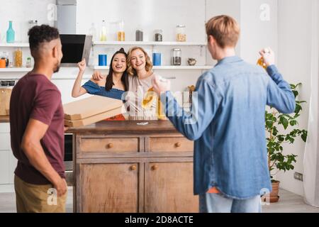 Rückansicht von multikulturellen Freunden, die Pizzaboxen und Bier halten In der Nähe Frauen zeigen yeah Geste Stockfoto