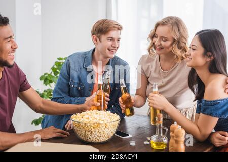 Aufgeregt multikulturelle Freunde klirren Flaschen Bier in der Nähe von Popcorn während Party Stockfoto