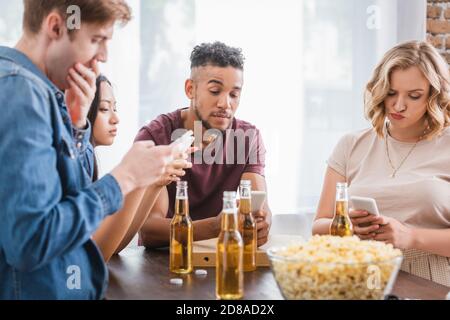Selektiver Fokus von multiethnischen Freunden chatten auf Mobiltelefonen in der Nähe Bier und Popcorn während der Party Stockfoto