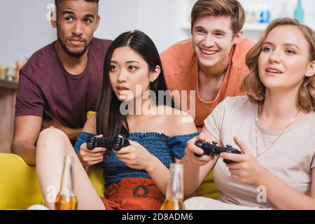 KIEW, UKRAINE - 28. JULI 2020: Aufgeregte Frauen spielen Videospiel in der Nähe von multikulturellen Freunden während der Party Stockfoto