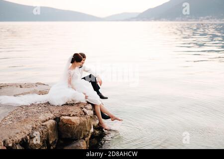 Die Braut und der Bräutigam sitzen auf dem Pier und Braut Schwingt ihre Füße im Wasser Stockfoto