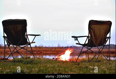 Ein Paar Touristenstühle, ein Klappstuhl, ein Lagerfeuer am Flussufer am späten Abend, Dämmerung im Herbst bei Sonnenuntergang, Entspannung und Ruhe, die b Stockfoto