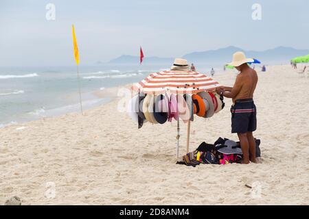 BARRA da TIJUCA, RIO DE JANEIRO, BRASILIEN - 2. JANUAR 2020: Strandverkäufer Mann steht am Strand und bereitet seine Hüte zum Verkauf vor. Stockfoto