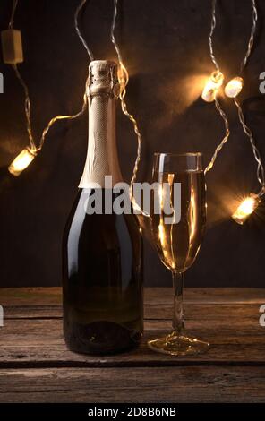 Weihnachtlicher Hintergrund mit einer Flasche Champagner und einem vollen Glas auf dem Hintergrund einer glühenden Girlande im Dunkeln. Stockfoto
