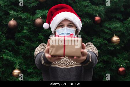 Corona Weihnachten Herzlichen Glückwunsch. Portrait Mann trägt santa Hut und Pullover in medizinischen Maske, Geschenk Geschenkbox mit rotem Band, weihnachtsbaum Stockfoto