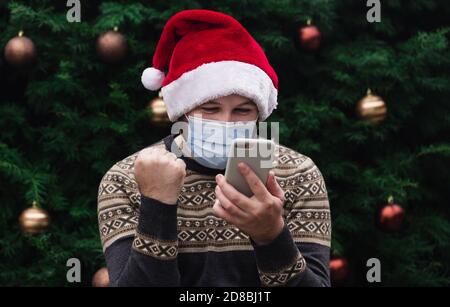 Weihnachten online Herzlichen Glückwunsch. Fröhlicher Mann trägt santa Hut und medizinische Maske im Gespräch am Telefon mit Freunden und Eltern. weihnachtsbaum Bokeh auf Stockfoto