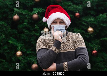 Weihnachtsidee. Nahaufnahme Porträt des Mannes trägt einen weihnachtsmann Hut, Weihnachten Pullover und medizinische Maske mit Emotion. Vor dem Hintergrund eines Weihnachtsfestes Stockfoto