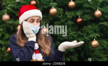 Weihnachten ohne Geschenk. Nahaufnahme Porträt einer Frau, die einen weihnachtsmann Hut und medizinische Maske mit Emotionen trägt. Vor dem Hintergrund eines Weihnachtsstr Stockfoto