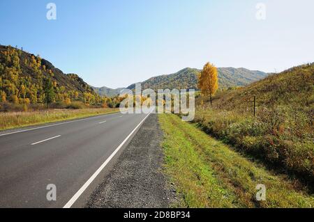 Eine gerade, zweispurige Asphaltstraße führt durch ein Herbsttal, umgeben von Hügeln und Bergen. Chuisky-Trakt, Altai, Sibirien, Russland. Stockfoto