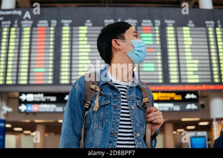 Asiatischer männlicher Reisender, der eine Gesichtsmaske trägt, die auf das Einsteigen in ein Flugzeug wartet. Junge Touristen warten auf Flughafen mit Abflug Bord im Hintergrund während Covid-19 Virus Pandemie Stockfoto