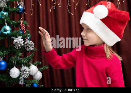 Niedliches kaukasisches fröhliches Mädchen in einem roten Pullover und einer Weihnachtsmütze ziert einen künstlichen Baum. Frohes neues Jahr und Weihnachtskonzept Stockfoto