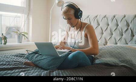 Junge kaukasische blonde Frau mit Headset spielen Spiel auf dem Laptop in ihrem Schlafzimmer. Hochwertige Fotos Stockfoto