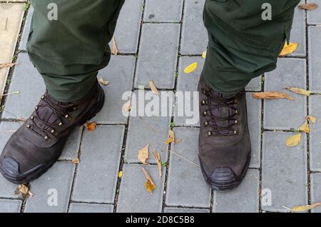 Herrenfüße in Militärstiefeln und Uniform stehen auf dem Bürgersteig inmitten gefallener Blätter. Soldat in braunen Stiefeln und grüner Uniform. Blick von oben auf eine A Stockfoto