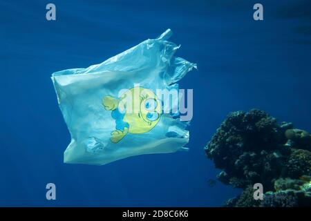 Plastikverschmutzung, Nahaufnahme einer weißen Plastiktüte mit gelbem Smiley, die langsam unter Wasser über das Korallenriff driftet Stockfoto