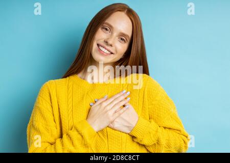 Schöne dankbar freundlich aussehende Rothaarige kaukasische Frau mit charmanten Lächeln in gelben Pullover halten beide Hände auf der Brust, zum Ausdruck Dankbarkeit, sh Stockfoto